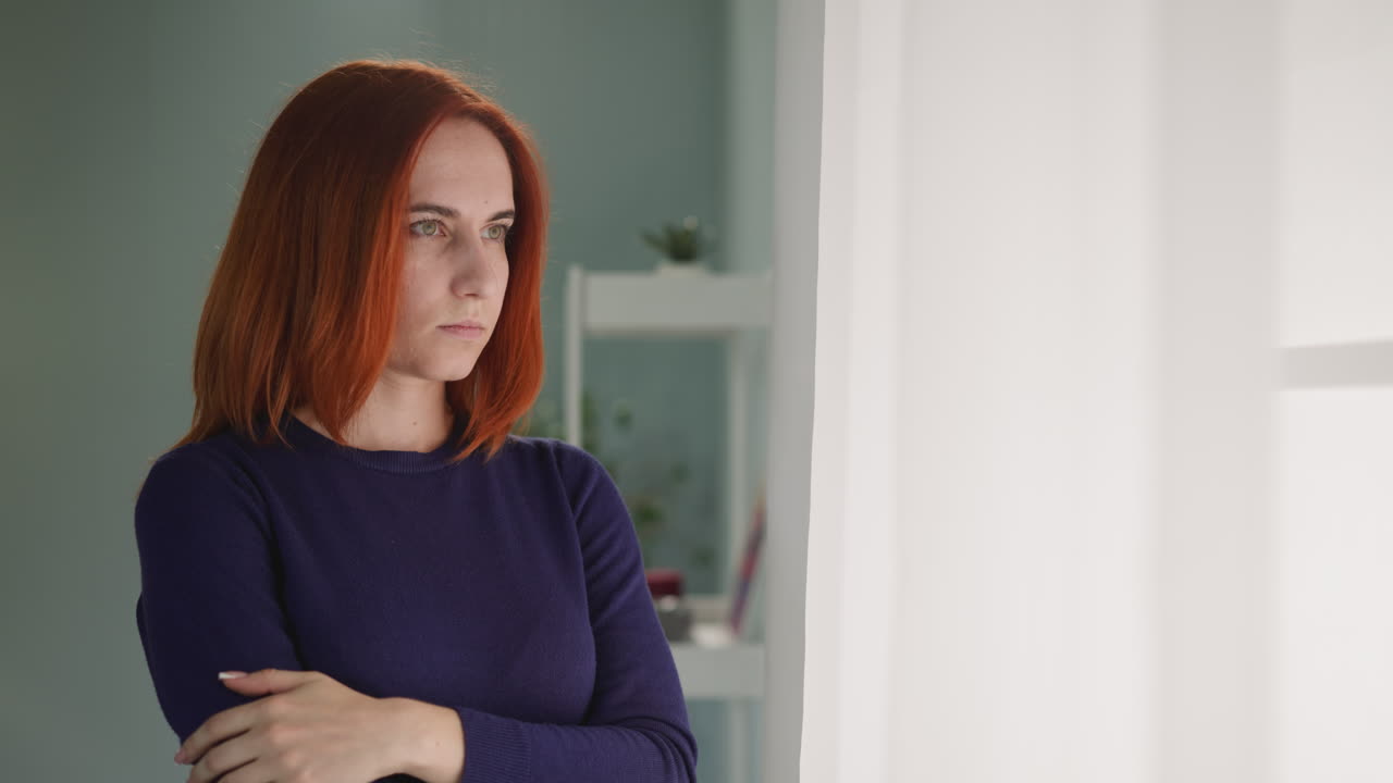 Mulher ruiva deprimida olha para fora pela janela iluminada.