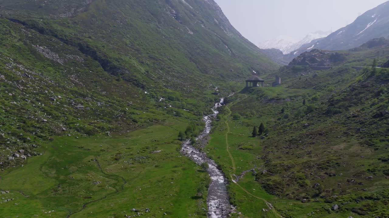 Veduta aerea dall'alto di un ruscello che serpeggia attraverso un'ampia valle verde incorniciata da pendii alpini rocciosi