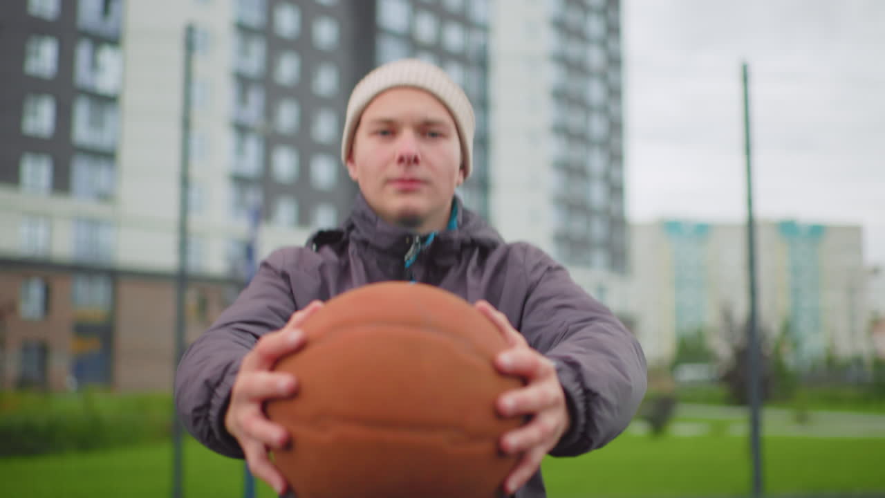 Primo piano di un giovane che tiene un pallone da basket in un campo urbano, mani che porgono il pallone verso la fotocamera, cielo nuvoloso, berretto e giacca invernale, palazzi e recinzione sfocati sullo sfondo, atmosfera sportiva di strada spontanea