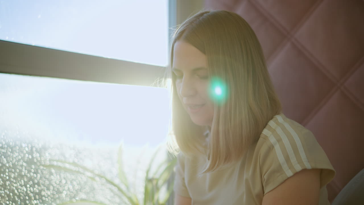 Young woman with long blonde hair sitting indoors near window sipping warm drink with whipped cream, enjoying relaxing moment in natural sunlight, holding cup with both hands in peaceful cozy atmosphere