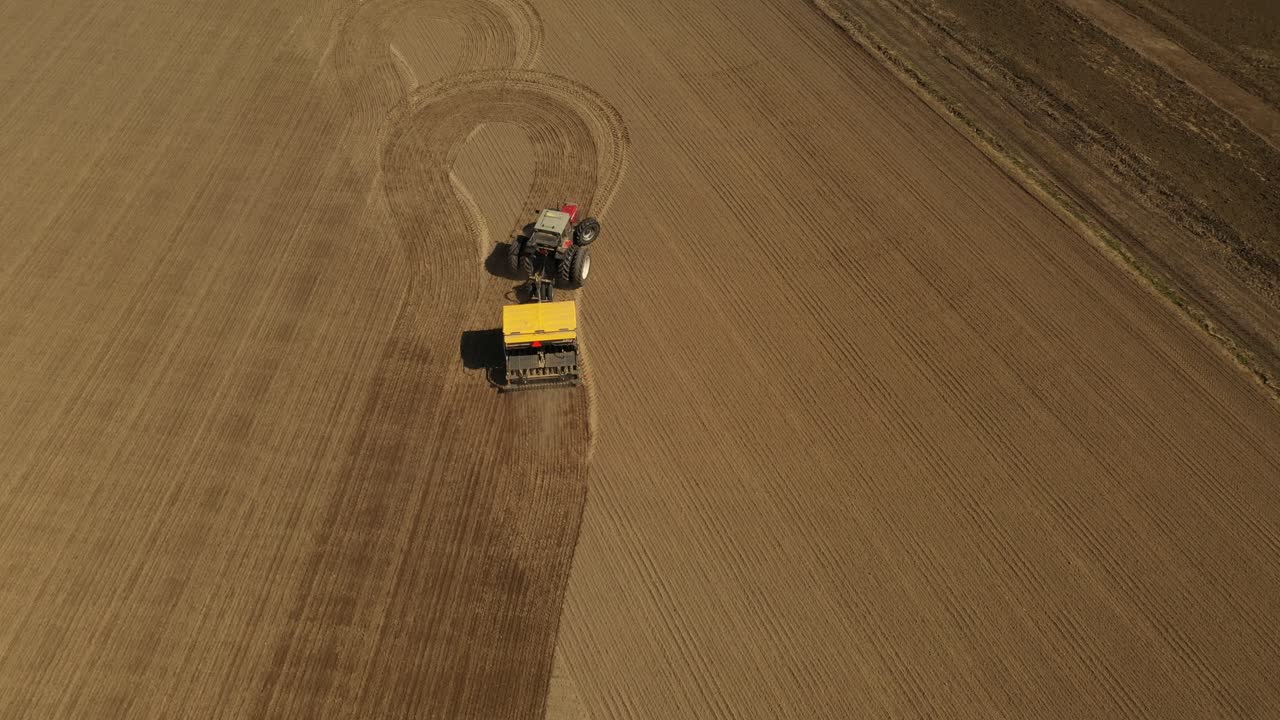 seguimiento de la vista aérea de un tractor rojo de dos ruedas sembrando semillas en un campo agrícola con una sembradora amarilla durante la primavera