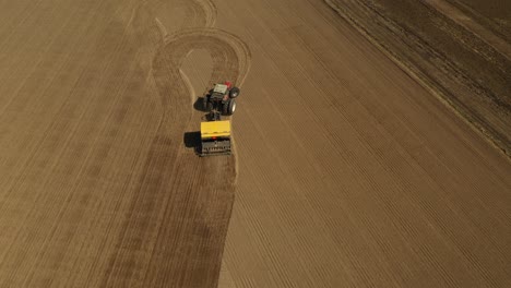 seguimiento de la vista aérea de un tractor rojo de dos ruedas sembrando semillas en un campo agrícola con una sembradora amarilla durante la primavera