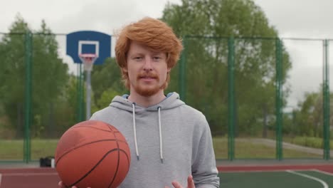 Joven-Atleta-Pelirrojo-Sonriente-Con-Sudadera-Sosteniendo-La-Pelota-Para-Jugar-Baloncesto-En-La-Cancha-De-Baloncesto