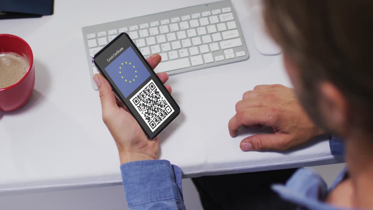 Man at desk holding smartphone with covid vaccination certificate, eu flag and qr code on screen