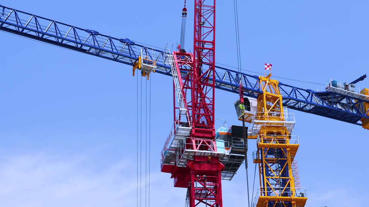 Two large tower cranes coordinate to lift and position a steel structure against a clear blue sky, with visible workers and dynamic camera panning