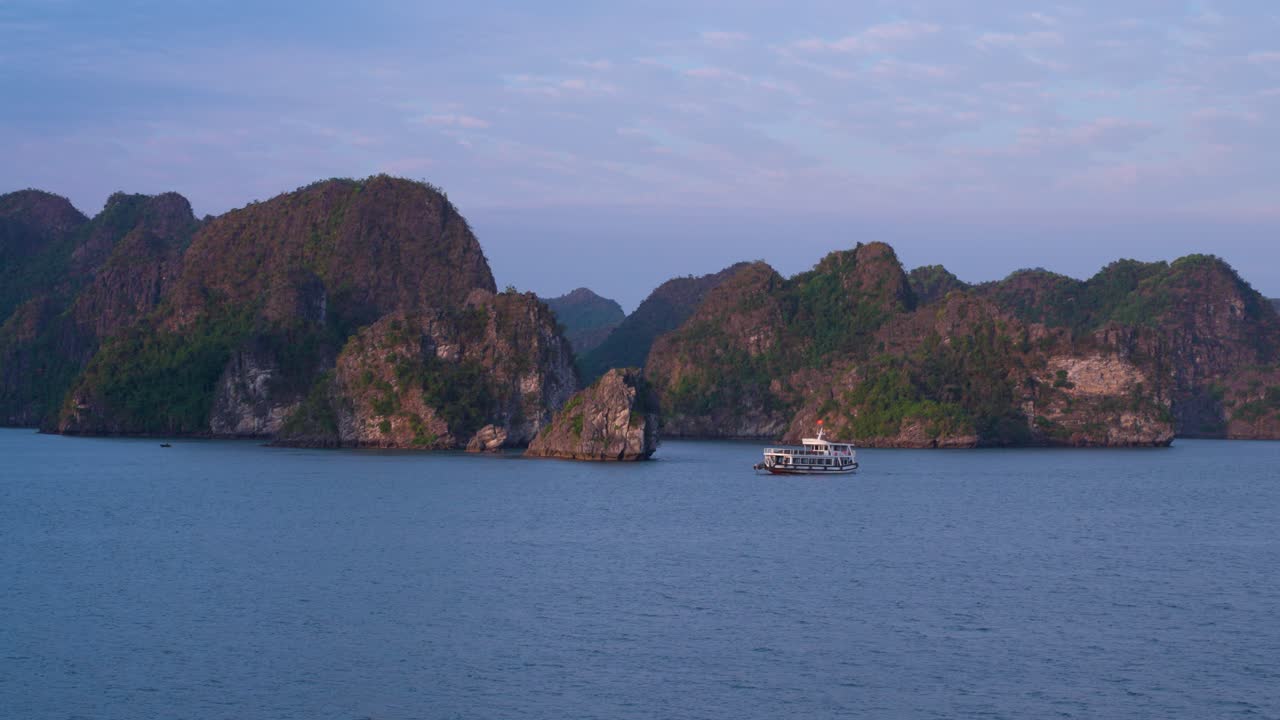 A traditional wooden cruise boat glides through the tranquil waters of Ha Long Bay, Vietnam, surrounded by towering limestone karsts and emerald-clad islands under a serene purple-blue twilight sky