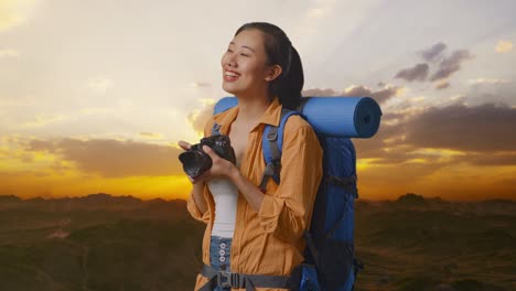 side view of asian female hiker with mountaineering backpack smiling and holding a camera in her hands then looking around while standing on the top of mountain during sunset time