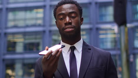 Young-Businessman-Wearing-Suit-Talking-On-Mobile-Phone-Using-Built-In-Microphone-Standing-Outside-Offices-In-The-Financial-District-Of-The-City-Of-London-UK-Shot-In-Real-Time-2