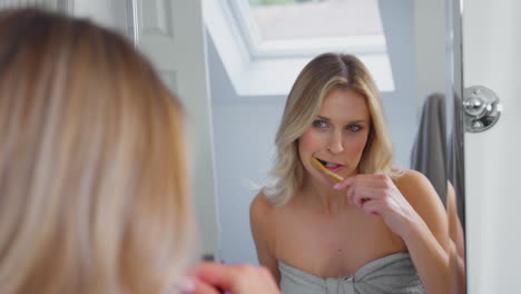 Mature-Woman-Brushing-Teeth-With-Wooden-Toothbrush-Looking-In-Mirror-In-Bathroom-At-Home