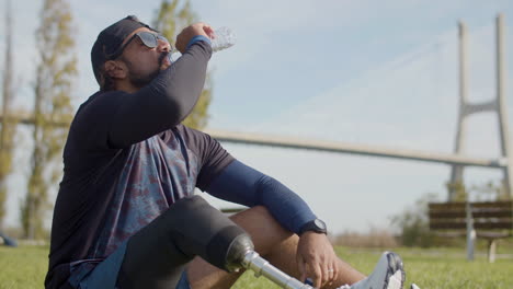 Plano-Medio-De-Un-Deportista-Cansado-Con-Una-Pierna-Artificial-Bebiendo-Agua-En-El-Césped-Del-Parque-Y-Relajándose-Después-De-Un-Intenso-Ejercicio-Cardiovascular-Por-La-Mañana