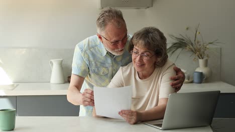 happy older married couple cuddle at kitchen discuss official papers