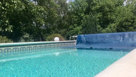 Rolling-up-solar-cover-reel-blanket-and-revealing-clean-swimming-pool-with-greenery-background