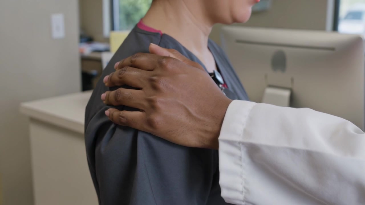Mano in camice bianco che si appoggia sulla spalla sinistra di un membro del personale al bancone della reception di una clinica, con uno scrub grigio