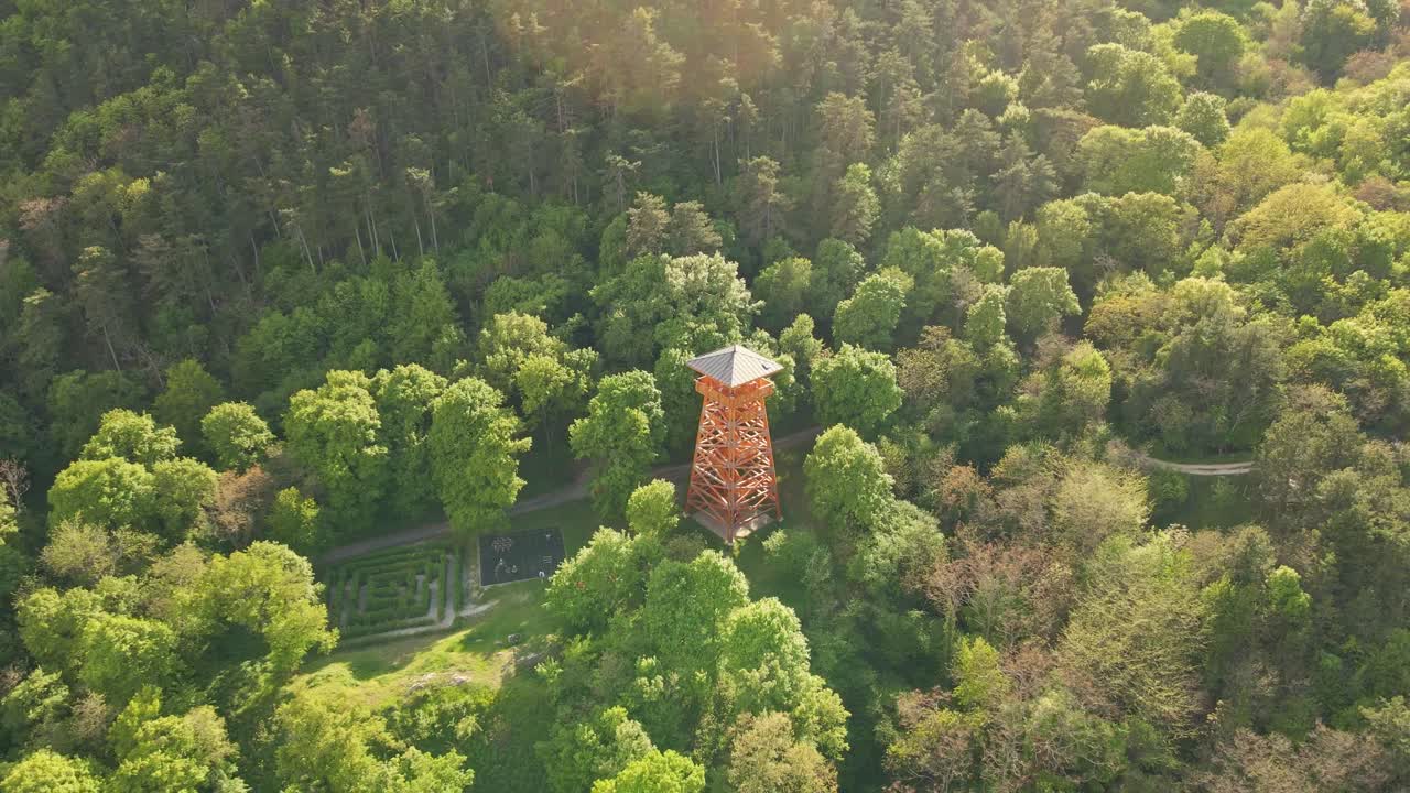 An aerial video of a wooden observation tower rising above a lush green forest, offering panoramic views of the surrounding countryside. Fly from the tower with a drone.