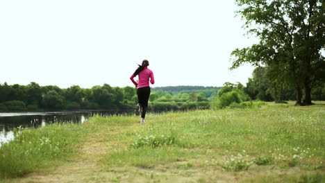 Brunette-with-long-hair-in-headphones-runs-along-the-river-in-the-Park-in-the-morning-at-sunrise-in-the-summer-in-a-pink-jacket-and-black-pants