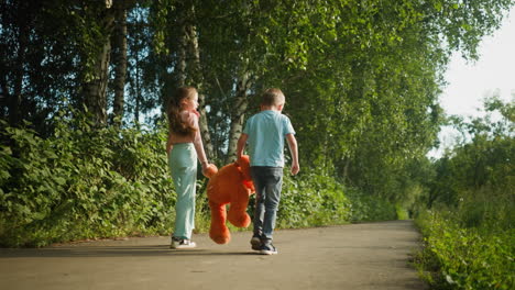 back view of two children walking side by side on quiet forest path holding large orange teddy bear, surrounded by lush trees and greenery, basking in soft daylight