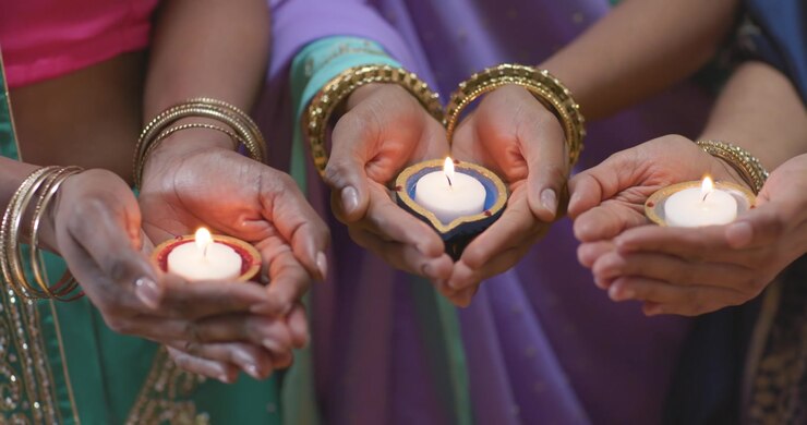 Three cupped hands are holding small lit lamps, steadying and aligning them during ritual offering