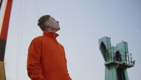 Hübscher-Junger-Containerlagerarbeiter-In-Orangefarbener-Uniform,-Der-Neben-Dem-Schiff-Im-Hafen-Steht