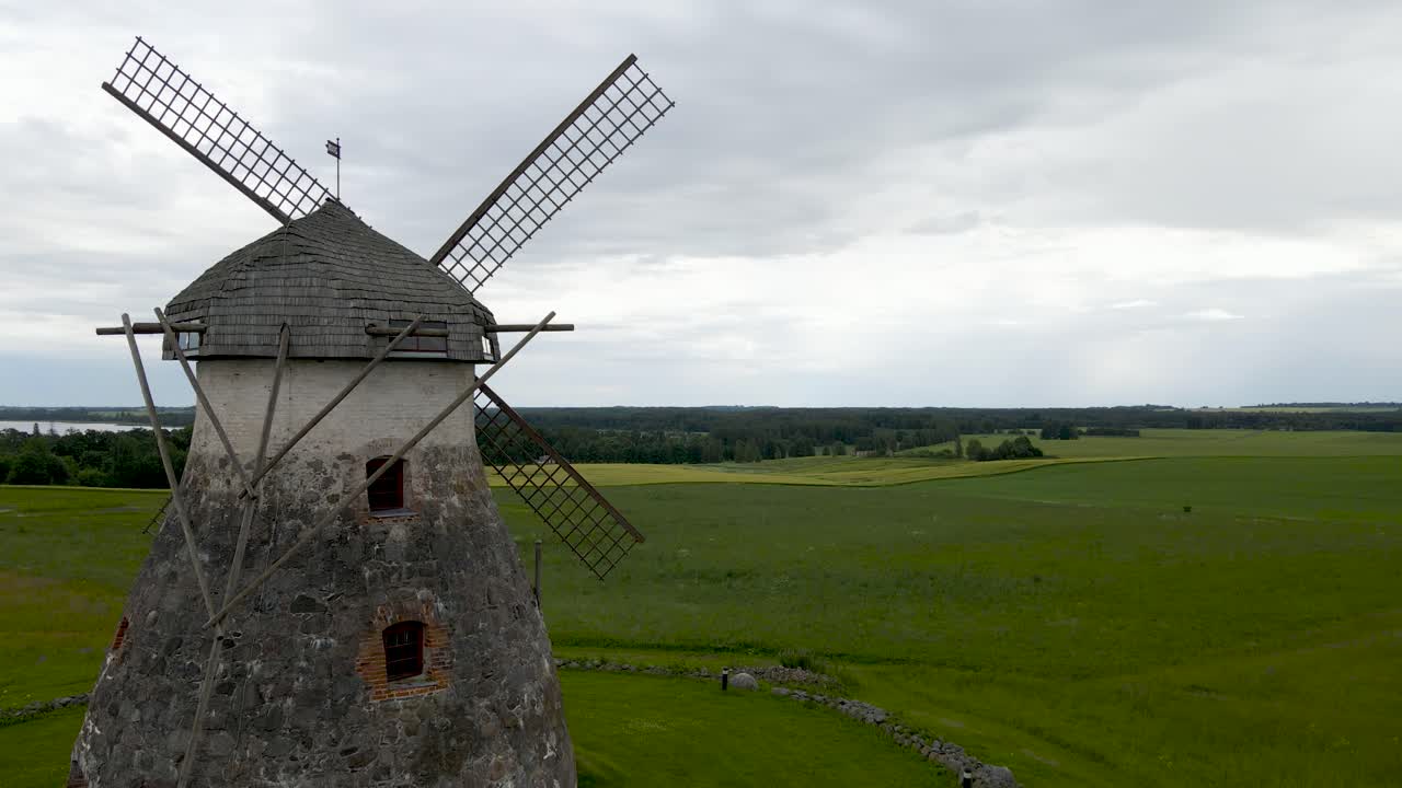 Gorgeous aerial drone footage ascending and showing a large and old vintage stone windmill in Kuremaa Estonia during a cloudy summer day with horizon visible at the background. Large flaps on the mill