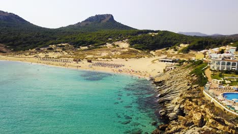 Approaching-drone-shot-of-Cala-Mesquida,-moving-above-some-luxurious-hotels-on-the-right-side,-above-the-cliffs-at-the-beachfront-on-t-he-island-of-Mallorca,-Spain