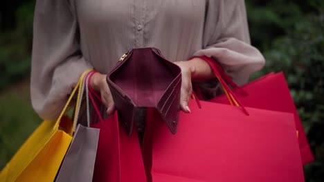 unrecognizable caucasian senior woman opening empty wallet showing no money inside. wrinkled female hands with shopping bags and billfold standing outdoors in park. spending and sales concept.