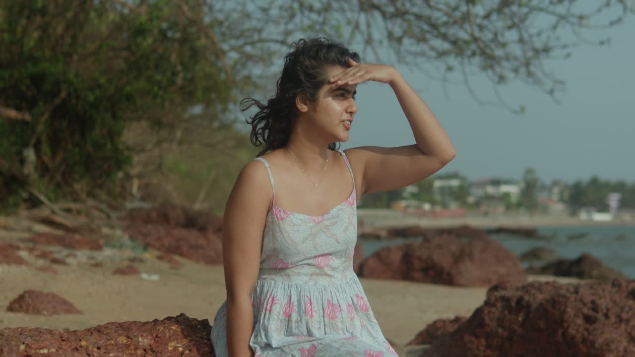 A young indian female is sitting on a rock by the water, looking out at the ocean