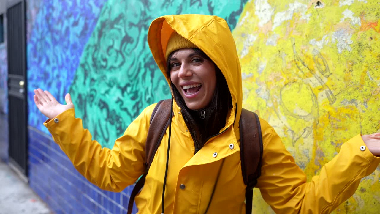 Glückliche junge Frau in gelbem Regenmantel lächelt vor einer bunten Wand.