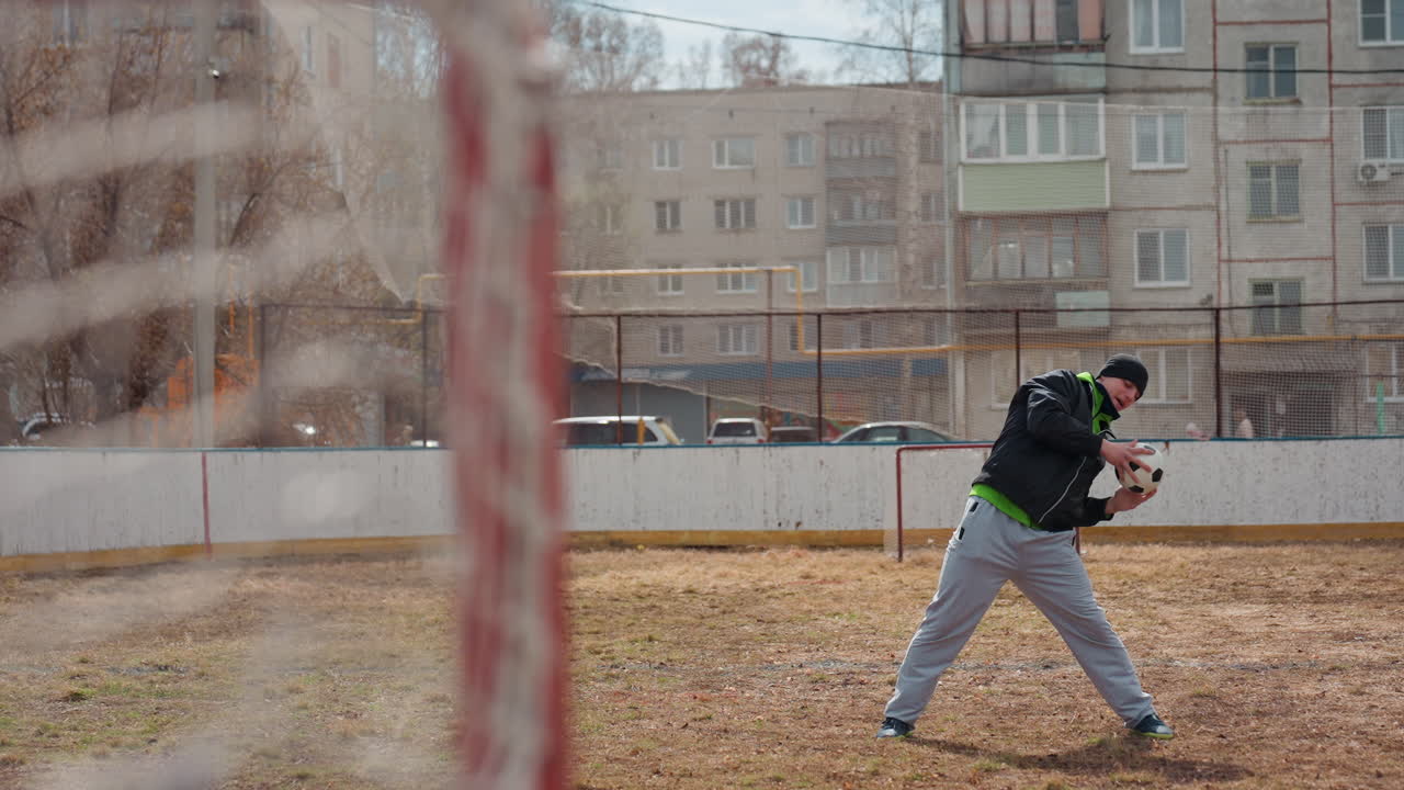 Un portero blanco practica lanzamientos y recepciones cerca de una portería pequeña, en una pista urbana vacía, con luz fría, postura concentrada y extensión del brazo, entrenando el reflejo y la coordinación, de forma metódica y repetitiva