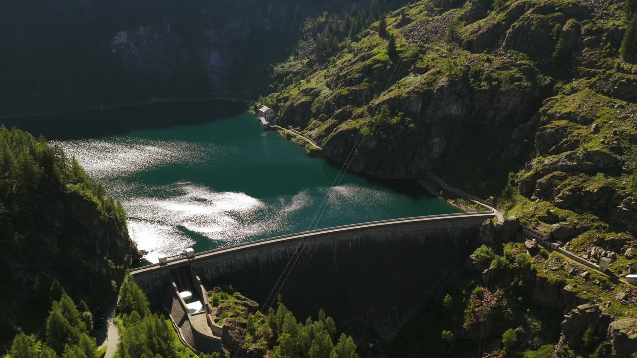Aerial view of Campliccioli Dam and reservoir in Valle Antrona. Sun reflects on green water surrounded by lush alpine mountains. For renewable energy or nature content, Italy