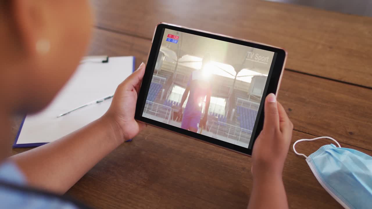 compuesto de una mujer sentada en una mesa con una máscara facial, viendo un evento deportivo en la tableta
