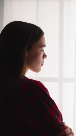 pretty teenage girl with loose hair in checkered shirt stands near french window in room zoom in slow motion. lady thinks and dreams spending time at home