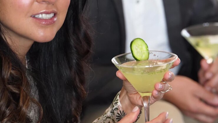 Donna tiene in mano un elegante Martini al cetriolo a un cocktail party mentre conversa con qualcuno che parla in un gruppo di persone. La bevanda o il drink misto è di colore verde con una guarnizione a fetta. Nessun volto