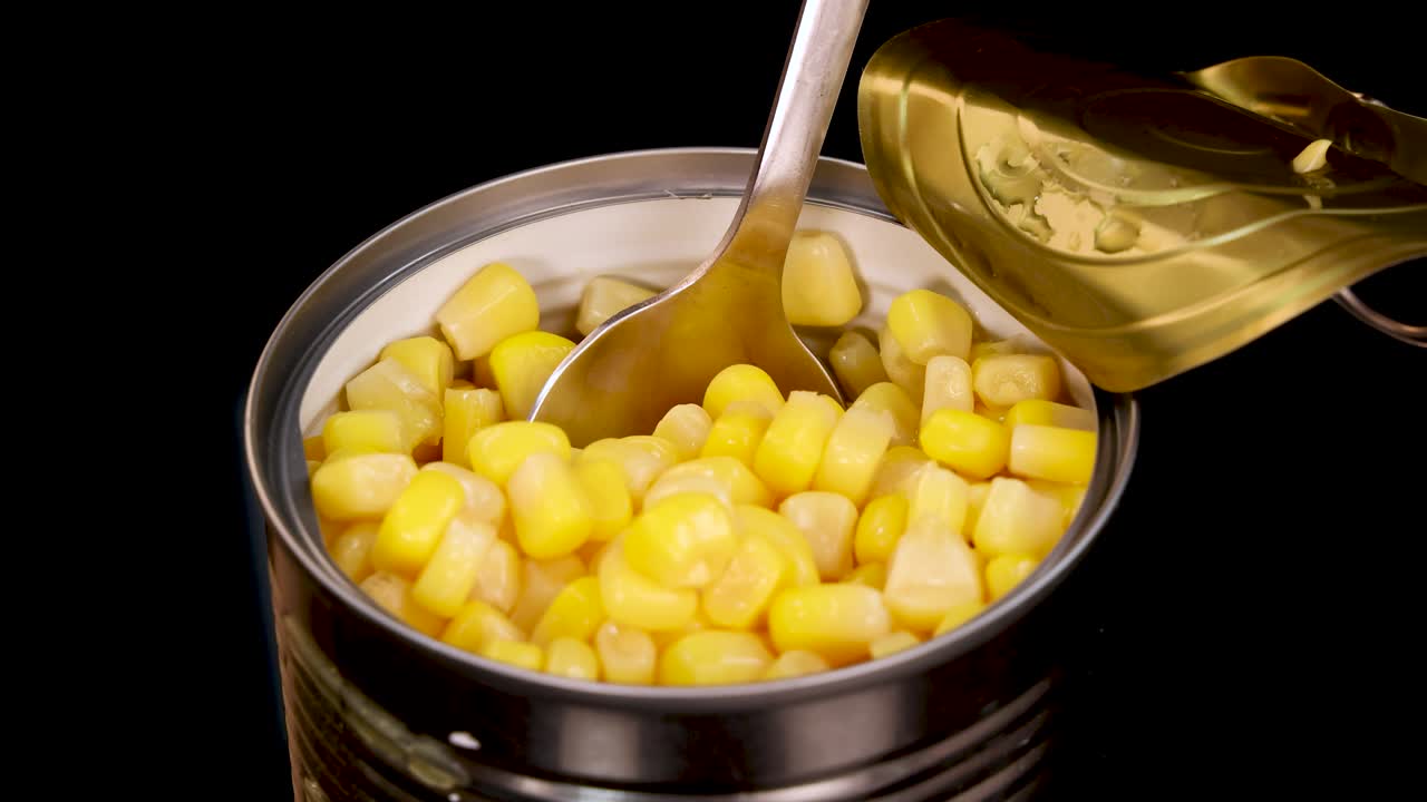 A metal spoon scoops yellow sweet corn kernels from an opened tin can, isolated on a black background with even, studio lighting and close-up framing