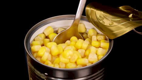 a metal spoon scoops yellow sweet corn kernels from an opened tin can, isolated on a black background with even, studio lighting and close-up framing