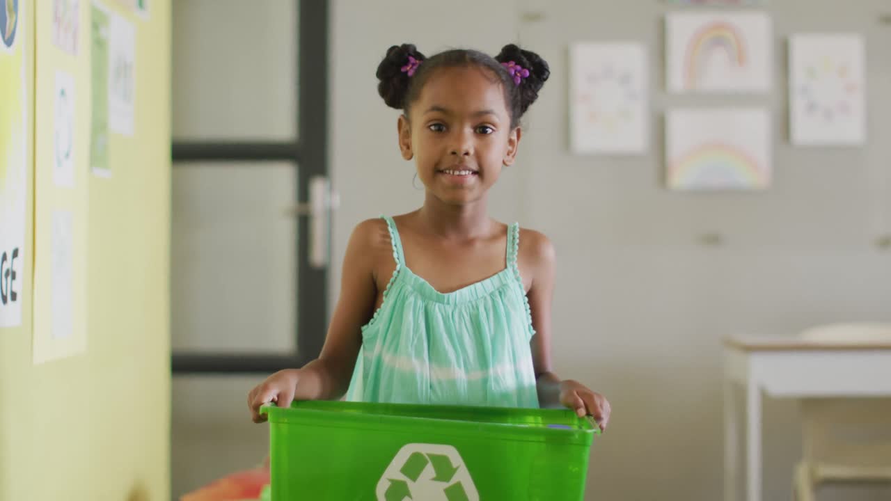 Video of happy african american girl holding box with recycling symbol in classroom