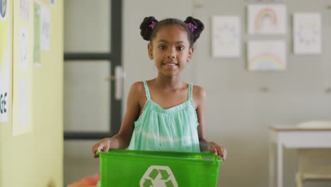 Video-Eines-Glücklichen-Afroamerikanischen-Mädchens,-Das-Eine-Box-Mit-Recycling-Symbol-Im-Klassenzimmer-Hält