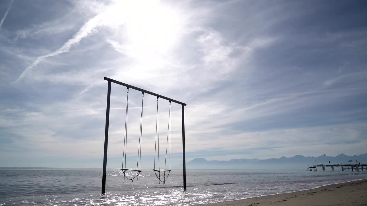 Swing set standing in shallow sea with bright sunburst sky gentle waves distant mountains and nearby pier creating calm seaside mood near Kemer in Antalya region