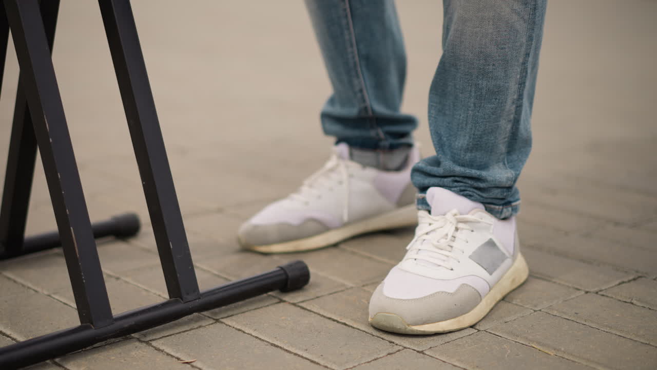 Sepatu kets jarak dekat di dekat stand keyboard, musisi (ras tidak diketahui) mengetuk kaki, ujung celana jeans denim, detail sepatu kets kasual di trotoar kota, nuansa pertunjukan jalanan langsung di luar ruangan dengan ritme dan gerakan halus.