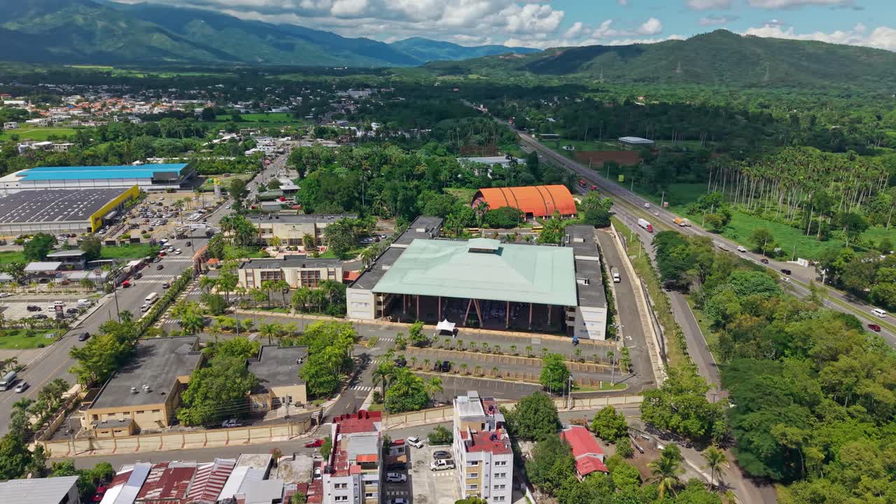 Autonomous University in Bonao with green mountains in background. Traffic on Main Street. Modern university infrastructure with pyramidal roof. Aerial orbit shot