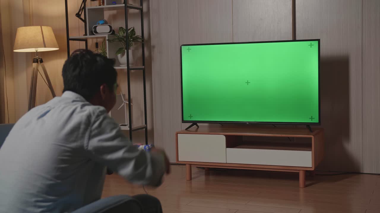 Un uomo è seduto sul divano e gioca a un videogioco con una TV a schermo verde, festeggiando la vittoria.