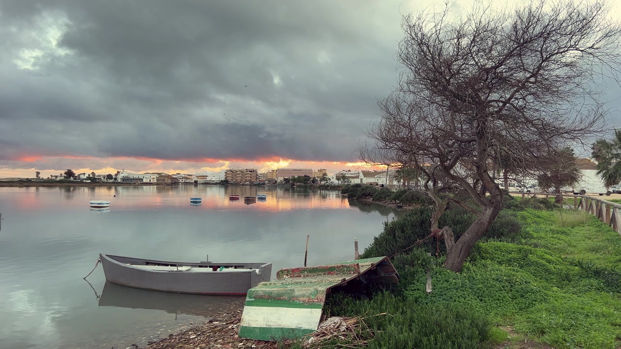 Scena tranquilla di una barca da pesca ormeggiata sul fiume Barbate a Barbate, Andalusia, Spagna, all'alba sotto un cielo nuvoloso e drammatico, con i riflessi della città e un'alba dai colori vivaci sulle acque tranquille.