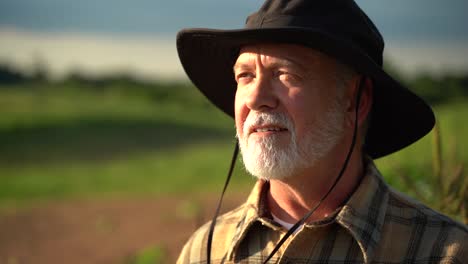 Close-up-portrait-of-senior-caucasian-good-looking-wise-man-farmer-in-a-hat-looking-at-the-side,-turning-face-to-the-camera-in-a-field