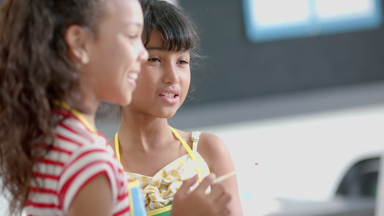 Two biracial girls talk in school classroom, one holding a pencil