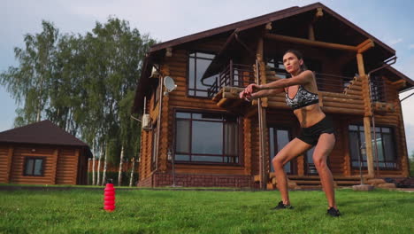 a slender young woman in black sportswear performs squats on the grass near her country house