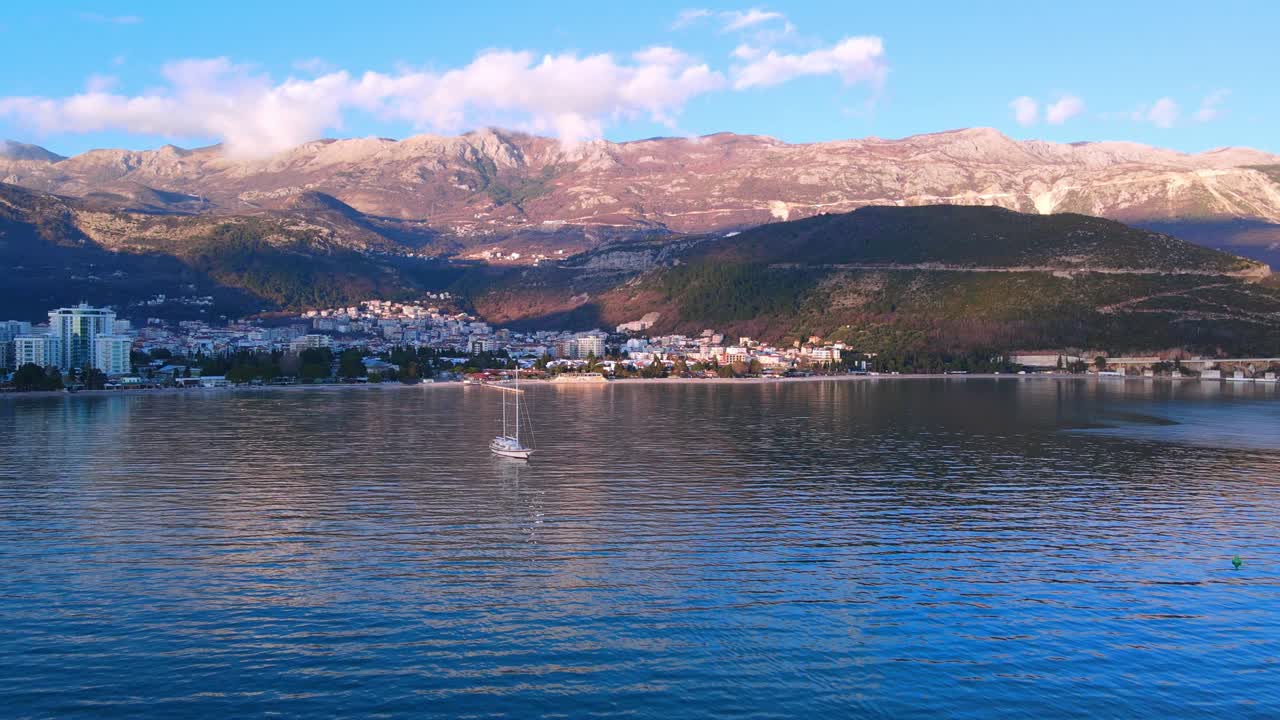 Deflated yacht on turquoise sea near tourist city against large mountains under orange cloudy sky in evening round aerial flight