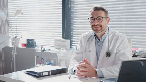 retrato de un médico de sexo masculino maduro sonriente con una bata blanca y un estetoscopio sentado detrás del escritorio en la oficina