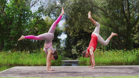 Caucasian-woman-and-man-practicing-yoga-outdoors,-standing-on-hands-on-wooden-jetty-near-forest