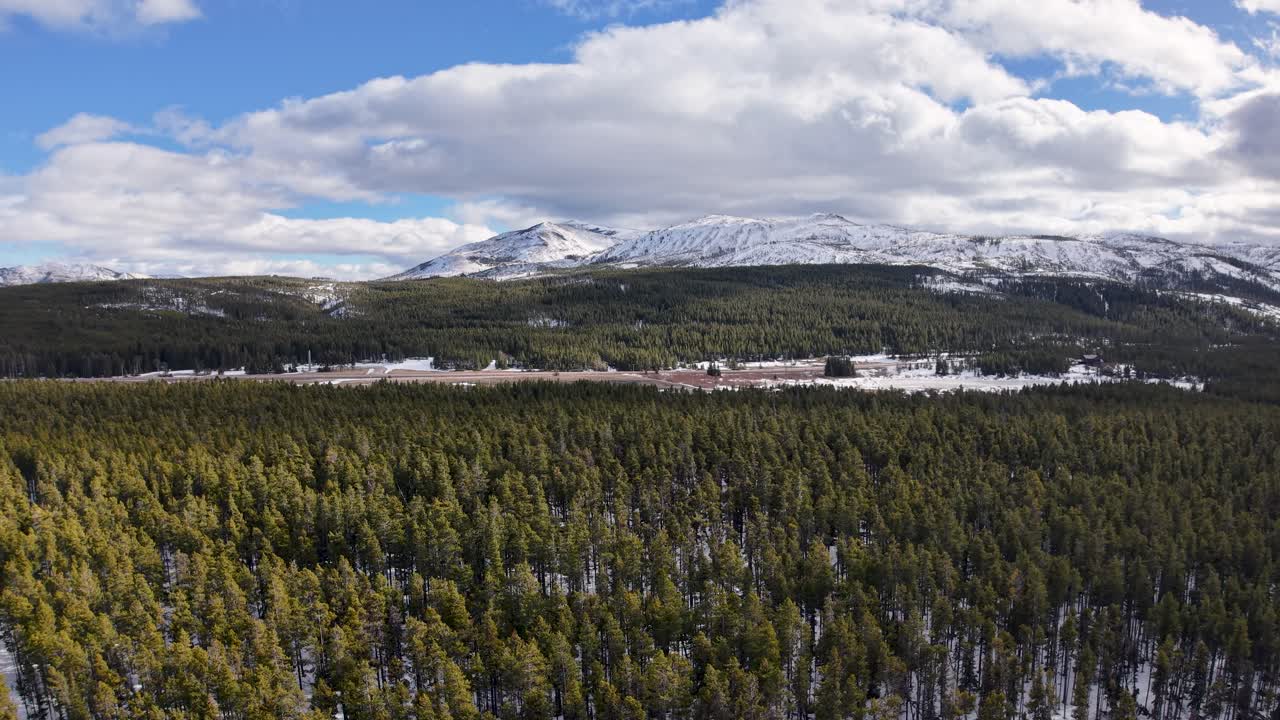 Il filmato del drone cattura un'ampia vista aerea di una foresta di conifere in Montana con montagne innevate e cielo parzialmente nuvoloso.