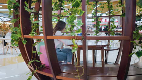 una joven adulta sentada en un asiento decorativo operando su teléfono, rodeada de luces decorativas brillantes, con gente bulliciosa en el fondo