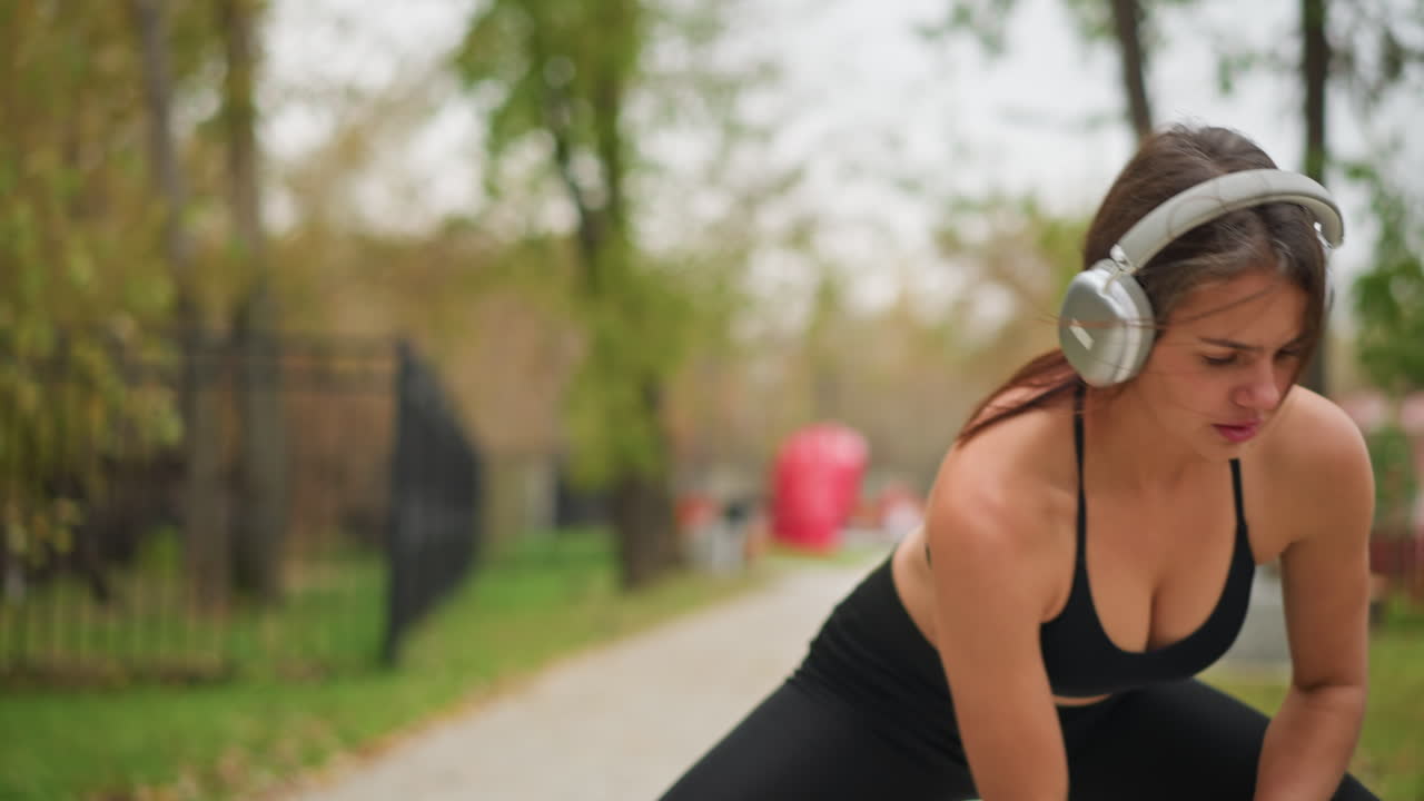 Portrait view of young woman in park outdoor training, stretching, and listening to music with headphones, blurred background with trees and playground, highlighting fitness and wellness lifestyle